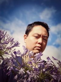 Low angle view of young man holding purple agapanthus lilies against cloudy blue sky.