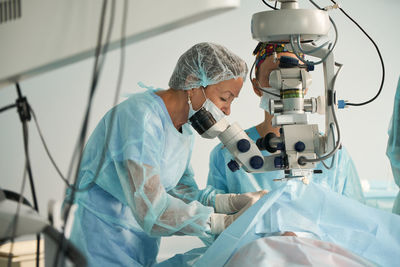 Two adult females doctor in sterile mask and ornamental medical cap looking through surgical microscope against crop coworker in hospital