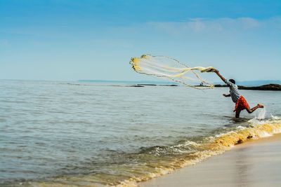 Full length of man throwing fishing net in sea against blue sky