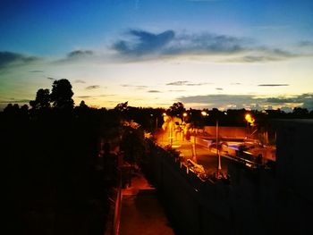 Illuminated road amidst silhouette trees against sky at sunset