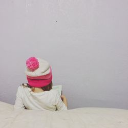 Rear view of girl in knit hat reading book against wall