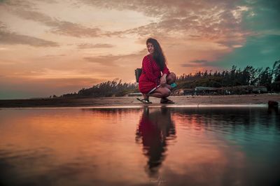 Woman sitting by lake against sky during sunset