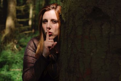 Portrait of young woman with finger on lips standing by tree in forest
