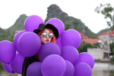 Portrait of young woman with dyed hair holding purple balloons while sitting against clear sky