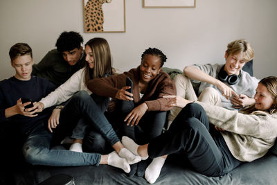 Smiling teenage friends using social media on mobile phones while relaxing on sofa at home