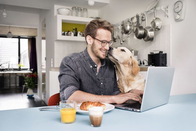 Smiling man with dog using laptop in kitchen at home