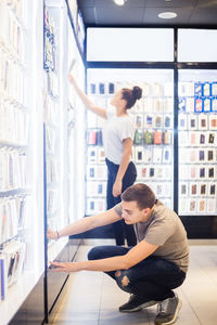 Full length of young male salesman crouching by illuminated rack while female customer shopping in mobile store
