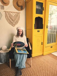 Young woman in beret sits in cozy home, room with music record in hands. vintage yellow cabinet.