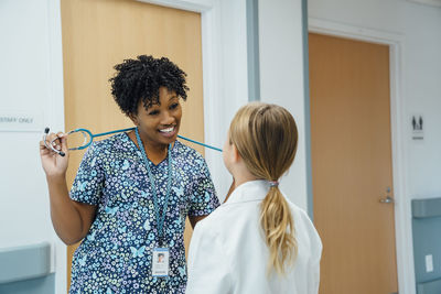 Cheerful pediatrician holding stethoscope while talking with girl in hospital corridor