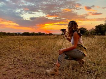 Woman looking through binoculars while crouching on land during sunset