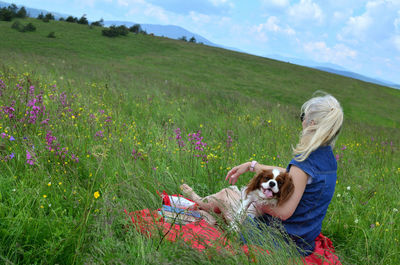 Blonde woman with her dog relaxing on a meadow surrounded with pink flowers and observing landscape