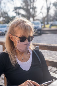 Mature adult blonde woman smiling with glasses wearing face mask texting to smartphone