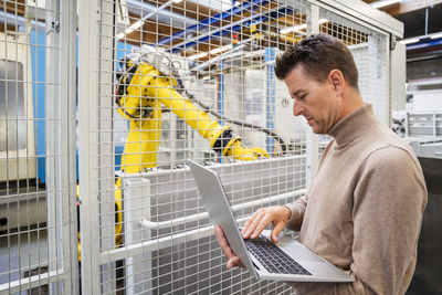 Mature businessman using laptop in front of robotic arm