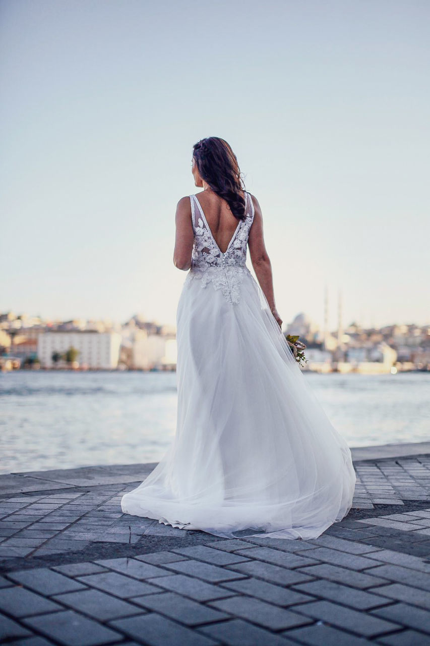 wedding dress, bride, adult, women, wedding, female, event, newlywed, young adult, one person, celebration, gown, dress, bridal clothing, fashion, clothing, full length, sky, architecture, nature, blue, emotion, standing, life events, city, married, love, white, positive emotion, ceremony, copy space, rear view, outdoors, formal wear, day, water, hairstyle, lifestyles, sunlight, happiness, brown hair, elegance, beginnings, looking, building exterior, person, clear sky, veil, portrait