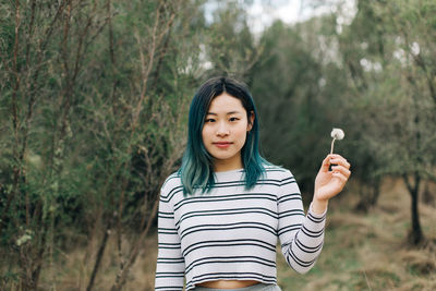 Portrait of young woman holding dandelion while standing against plants