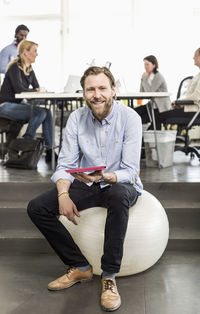 Portrait of mid adult businessman with digital tablet sitting on fitness ball in office