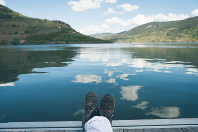 Low section of man resting on pier over lake by mountains against sky