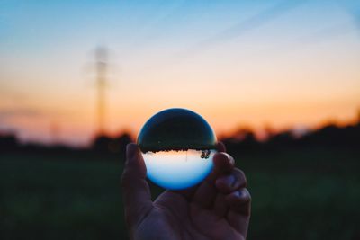 Close-up of human hand holding crystal ball against sky during sunset