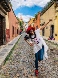 Portrait of cheerful woman holding camera while standing on footpath in town