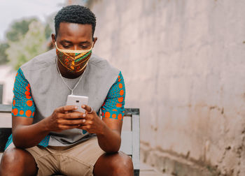 Young man using mobile phone while sitting against wall