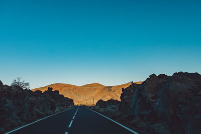 Road leading towards mountains against clear blue sky