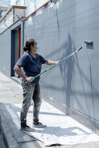 Side view of professional female painter in protective mask standing on sidewalk and painting wall using roller on city street