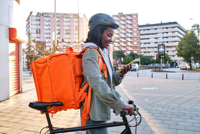 Side view of positive african american female cyclist with thermo backpack standing on paved sidewalk while checking order information on cellphone