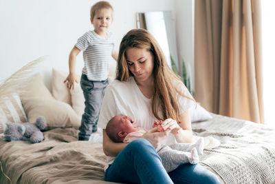 Young caucasian mother with newborn baby and toddler son on bed at home.