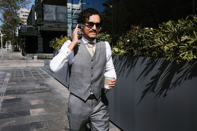 Positive bearded male entrepreneur in elegant suit having phone conversation on smartphone and looking away while standing with takeaway coffee on the street with sunglasses