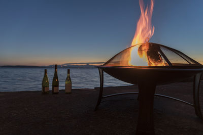 Wine bottles by burning fire pit at beach against sky during sunset