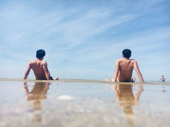 Rear view of shirtless men relaxing at beach against sky