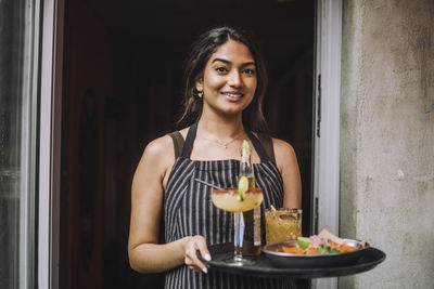 Portrait of smiling waitress holding tray of food and drinks at bar doorway