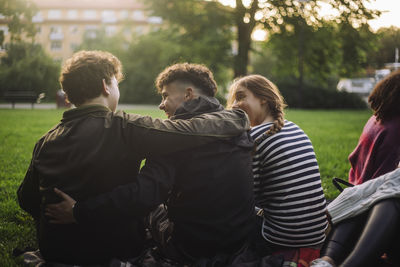 Rear view of male and female friends sitting with arms around at park