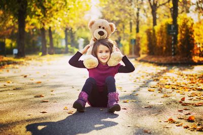 Full length portrait of smiling girl with teddy bear sitting on road at park during autumn