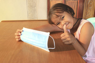 Portrait of cute girl holding protective face mask and showing thumbs up on table at home