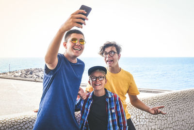 Smiling friends taking selfie using smart phone against sea