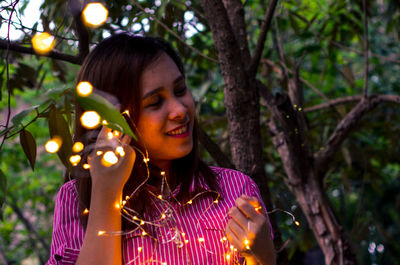 Woman with illuminated string light against trees