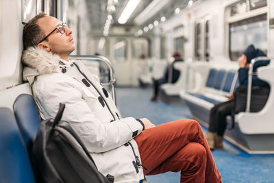 Man with glasses in white jacket and maroon trousers fell asleep on subway wagon on his way home.