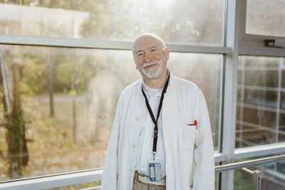 Portrait of smiling senior male doctor leaning on railing against window at hospital corridor