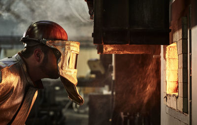 Blue-collar worker wearing protective helmet looking at burning furnace in industry