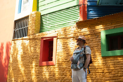 Rear view of man standing against bright colourful building with newborn baby 
