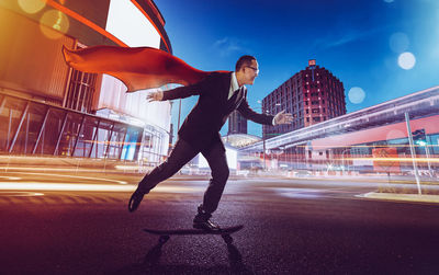 Side view of businessman wearing cape skateboarding on city street at night