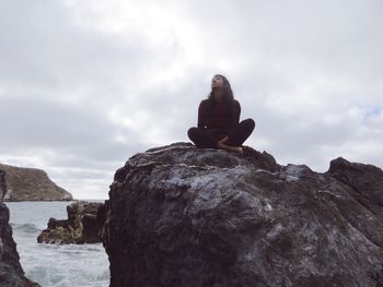 Woman sitting on rock against cloudy sky