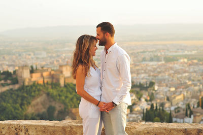 Side view of young romantic couple standing by retaining wall against city