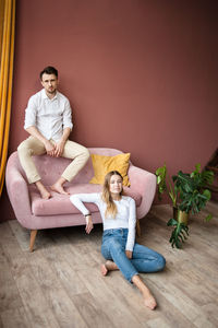 Man sits on pink sofa's back against wall, woman sits on floor near.
