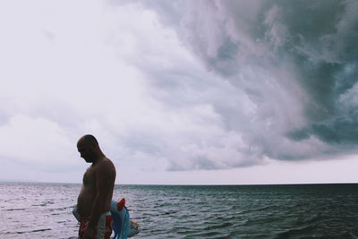 Shirtless mid adult man standing in sea against cloudy sky