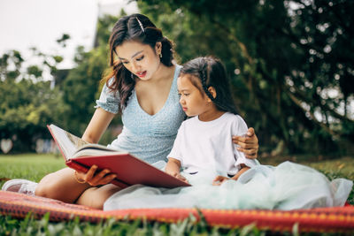 Smiling teacher and girl reading book sitting at park