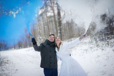 Smiling wedding couple holding distress flares while standing at forest during winter