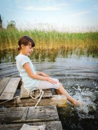 Little girl is sitting on a pier and splashing her legs on the lake. hot summer.