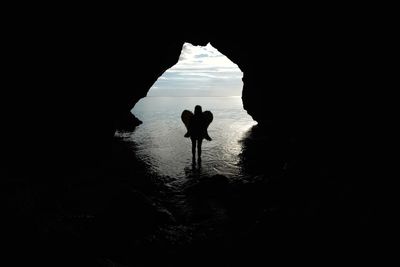 Silhouette woman wings standing in sea seen through cave